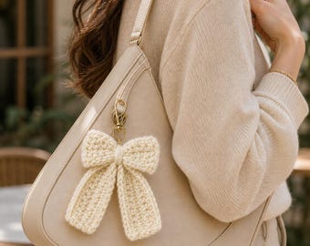 May include: A cream-colored crochet bow bag charm attached to a light beige shoulder bag with a gold-toned metal lobster clasp. The bow features a textured, hand-stitched yarn construction with two long, hanging tails and a central knot detail.