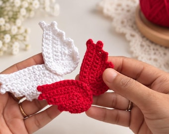 May include: Two crocheted angel wing appliques, one in solid white and one in solid red. Each wing features a textured, stitched pattern with scalloped edges along the top. The wings are separate pieces, crafted from yarn with a visible knit texture.