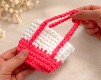 May include: A small, handmade crochet tote bag featuring a two-tone design with a bright neon pink base and handles, contrasted by a white upper section. The bag is constructed from thick, soft yarn with a visible textured stitch pattern throughout.
