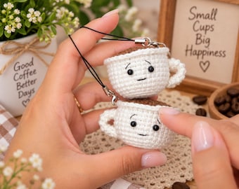 May include: Two small, white, crocheted coffee cup charms with brown tops, black bead eyes, and embroidered smiles. Each cup features a small handle and is attached to a thin black cord with a metal clasp for hanging as a decorative accessory.