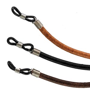 May include: Close-up of three eyeglass holders in brown, black, and dark brown leather. Each holder has a black loop for the glasses and silver metal coils at the connection point. The leather cords are round and smooth.