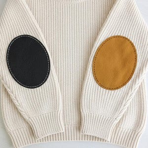 May include: A cream-coloured ribbed knit jumper featuring long sleeves with contrasting elbow patches. The left sleeve has a black oval patch with white stitching, while the right sleeve has a mustard yellow oval patch with matching stitching.