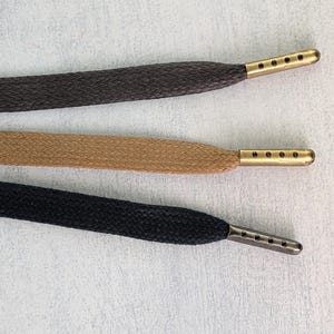 May include: Three shoelaces in shades of brown, tan, and black, each with a metal aglet. The aglets have multiple holes. The laces are flat and woven, showing a textured pattern. The background is a light grey.