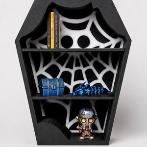Puede incluir: Estantería negra con forma de ataúd con tres estantes. El interior presenta un diseño de telaraña blanca. Los estantes contienen un cofre del tesoro azul, una figura de dragón azul, una figura de robot y dos libros.
