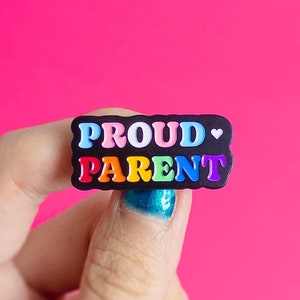 Puede incluir: Un botón negro con las palabras "PROUD PARENT" en colores arcoíris y un pequeño símbolo de corazón.