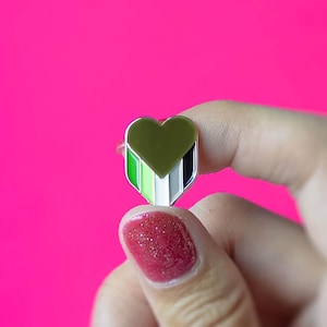 Op de afbeelding: Een groene, witte en zwarte hartvormige pin met een zilveren achterkant. De pin wordt vastgehouden door een hand met rode nagellak.