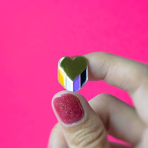 May include: A gold heart-shaped pin featuring the non-binary flag colours: yellow, white, purple, and black. The pin is held against a bright pink backdrop. A small accessory.