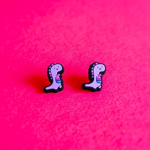 May include: A pair of purple dinosaur stud earrings on a pink background.