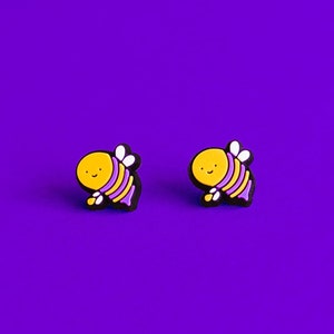 May include: A pair of small, yellow and purple bee earrings on a purple background.