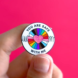 Può includere: Uno spillo in smalto bianco con un cerchio arcobaleno e un cuore rosa al centro. Il testo "You are safe with me" è scritto attorno al cerchio.