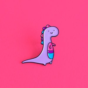 May include: Purple dinosaur enamel pin with pink, blue and white accents. The dinosaur has a smiling face and is standing on a pink background.