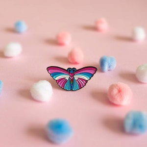 May include: A colourful butterfly pin with pink, blue and white wings. The pin is on a pink background with white, blue and pink pom-poms.