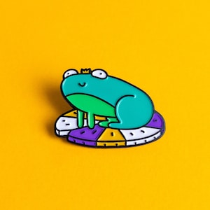 May include: A green frog wearing a crown sits on a purple, yellow and white patterned circle. The frog has a happy expression.