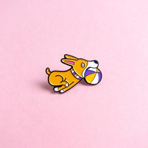 May include: A yellow and orange enamel pin of a cartoon dog running with a purple and yellow ball in its mouth.