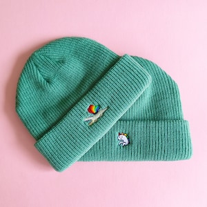 May include: Two green knit beanies with embroidered designs. One beanie has a rainbow unicorn and the other has a rainbow alligator with a rainbow balloon.