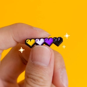 Op de afbeelding: Een zwarte emaille pin met vier gepixelde harten op een rij. De harten zijn geel, wit, paars en zwart.