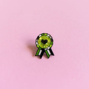 Puede incluir: Un pin de brillo verde, negro y blanco en forma de cinta con un corazón negro en el centro.