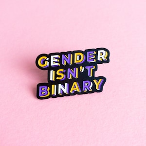 May include: A black enamel pin with the text "GENDER ISN'T BINARY" in yellow and purple letters. The pin is on a pink background.