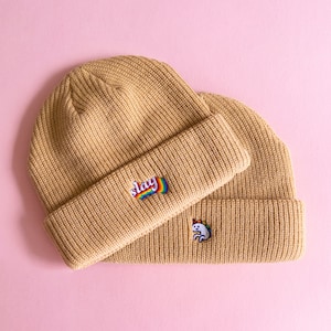 Peut inclure: Deux bonnets en tricot beige avec des motifs brodés. Le bonnet du dessus a un motif "slay" arc-en-ciel. Le bonnet du dessous a un motif de licorne blanche.