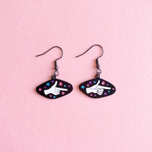 May include: A pair of black dangle earrings with a white hand pointing to the right. The hand is surrounded by pink and blue stars on a black background.