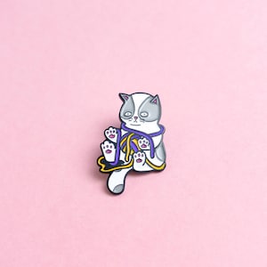 May include: An enamel pin featuring a white and grey cat with purple and yellow yarn. The cat is sitting and holding the yarn in its paws.