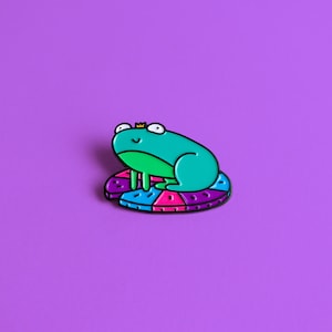 May include: A green frog wearing a gold crown sits on a colorful, multi-colored, circular cushion. The cushion is made of a soft, plush material and has a variety of colors, including purple, pink, blue, and green.