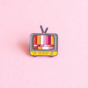 Pode incluir: Um broche de esmalte cinza e amarelo em forma de televisão vintage com uma tela estática colorida dentro. A tela está cheia de barras horizontais laranja, branco, rosa e vermelho.