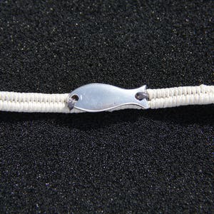 May include: A white cord bracelet with a silver fish charm. The bracelet is on a black background.