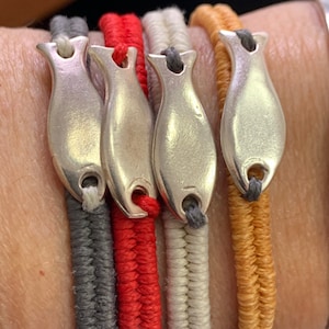 Könnte beinhalten: Vier geflochtene Armbänder mit silbernen Fisch-Anhängern. Die Armbänder sind in Grau, Rot, Weiß und Orange.