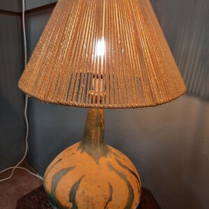 May include: A vintage table lamp featuring a woven rope lampshade and a ceramic base. The base has a bulbous shape with an orange and green design. The lamp is switched on, illuminating the interior. The lamp is a decorative lighting fixture.