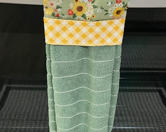 May include: A green kitchen towel with white stripes and a fringed edge, hanging from a metal bar. The top section features a floral pattern with sunflowers and a yellow gingham band, adding a touch of country charm to the kitchen.