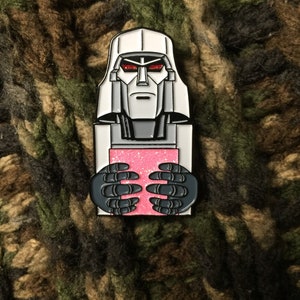 May include: A silver and black enamel pin featuring a cartoon depiction of Megatron, a character from the Transformers franchise. Megatron is holding a pink, glittery rectangle in his hands.