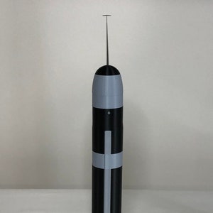 May include: A black and gray cylindrical tool with a thin, vertical metal rod extending from the top. The tool has gray bands and a cross-shaped design. The object is standing upright against a white background.