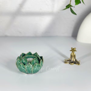 May include: A green ceramic lotus flower candle holder with gold accents and a small brass frog figurine in a yoga pose. The items are on a white surface with a blurred background.