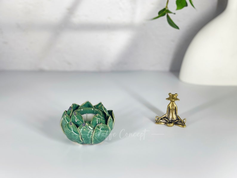 May include: A green ceramic lotus flower candle holder with gold accents and a small brass frog figurine in a yoga pose. The items are on a white surface with a blurred background.