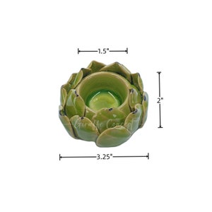 May include: A green ceramic artichoke-shaped candle holder. The candle holder is 8.26 cm wide, 5.08 cm tall, and 3.81 cm deep. The interior is a glossy green, and the exterior has a textured, leaf-like design. The text "Giraffe Concept" is visible.