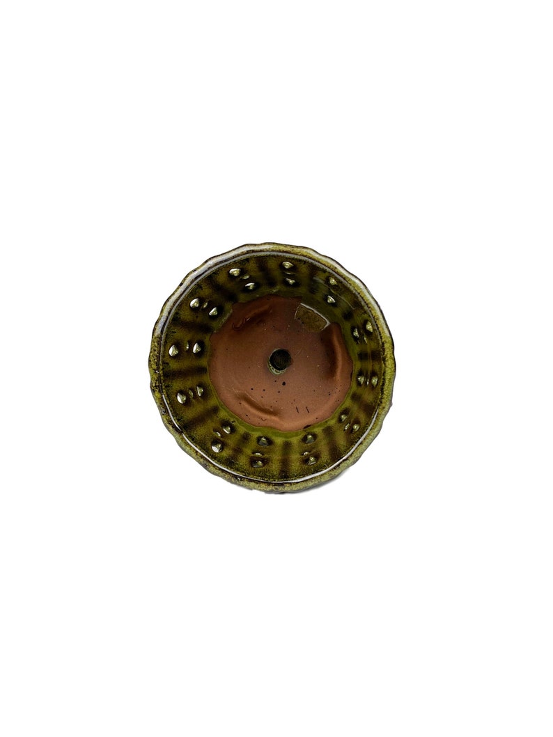 May include: The bottom of a green and brown ceramic pot with drainage holes. The pot has a textured surface and a slightly raised rim.