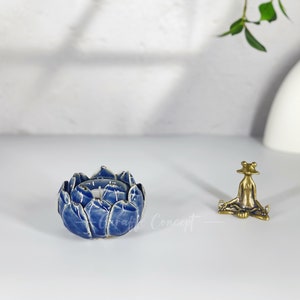May include: A blue ceramic lotus flower-shaped candle holder with white detailing. A small brass frog figurine is seated nearby. The items are on a white surface.
