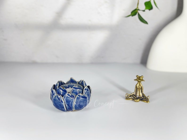 May include: A blue ceramic lotus flower-shaped candle holder with white detailing. A small brass frog figurine is seated nearby. The items are on a white surface.