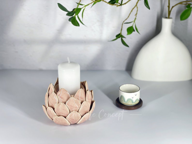 May include: A pink ceramic candle holder shaped like a flower with a white candle inside. The candle holder is on a white surface with a small white cup and saucer with a green and white design on a wooden coaster.