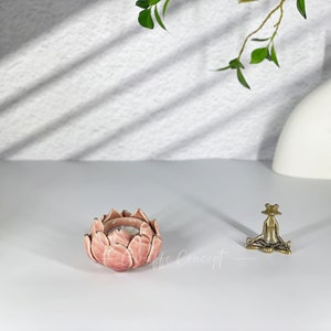 May include: A pink lotus flower-shaped candle holder with a lit candle inside. A small brass frog figurine is seated nearby. A white vase and greenery are in the background. The candle holder is approximately 10 cm in diameter.
