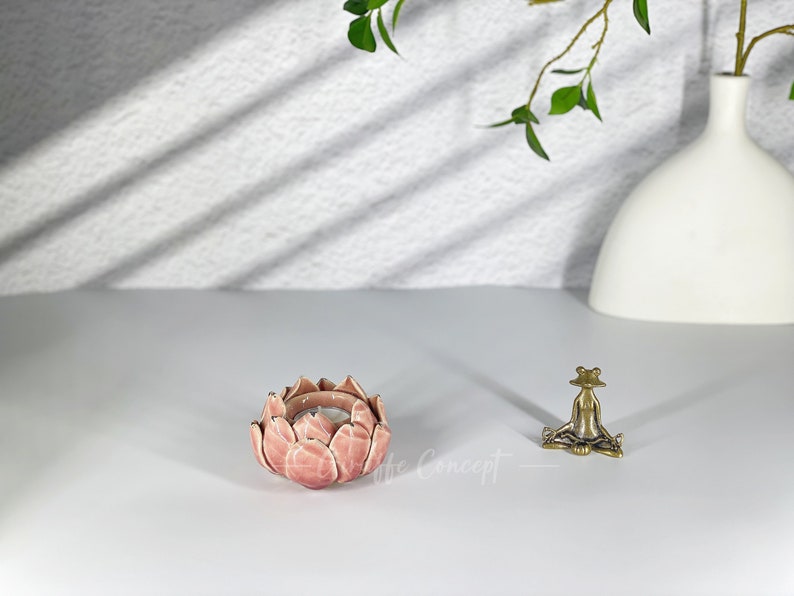 May include: A pink lotus flower-shaped candle holder with a lit candle inside. A small brass frog figurine is seated nearby. A white vase and greenery are in the background. The candle holder is approximately 10 cm in diameter.