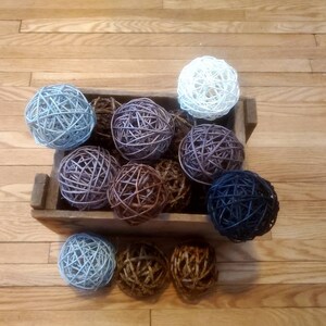 May include: A wooden crate filled with a variety of woven balls in shades of blue, brown, and purple. The balls are made of natural materials and have a rustic, handcrafted look.