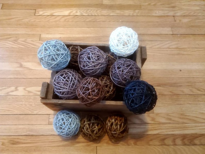 May include: A wooden crate filled with a variety of woven balls in shades of blue, brown, and purple. The balls are made of natural materials and have a rustic, handcrafted look.