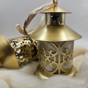 May include: Two gold metal lanterns with intricate cut-out designs. The lanterns are sitting on a white fluffy surface. One lantern has a star cut-out on the top and a white ribbon attached.