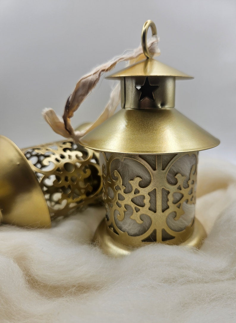 May include: Two gold metal lanterns with intricate cut-out designs. The lanterns are sitting on a white fluffy surface. One lantern has a star cut-out on the top and a white ribbon attached.
