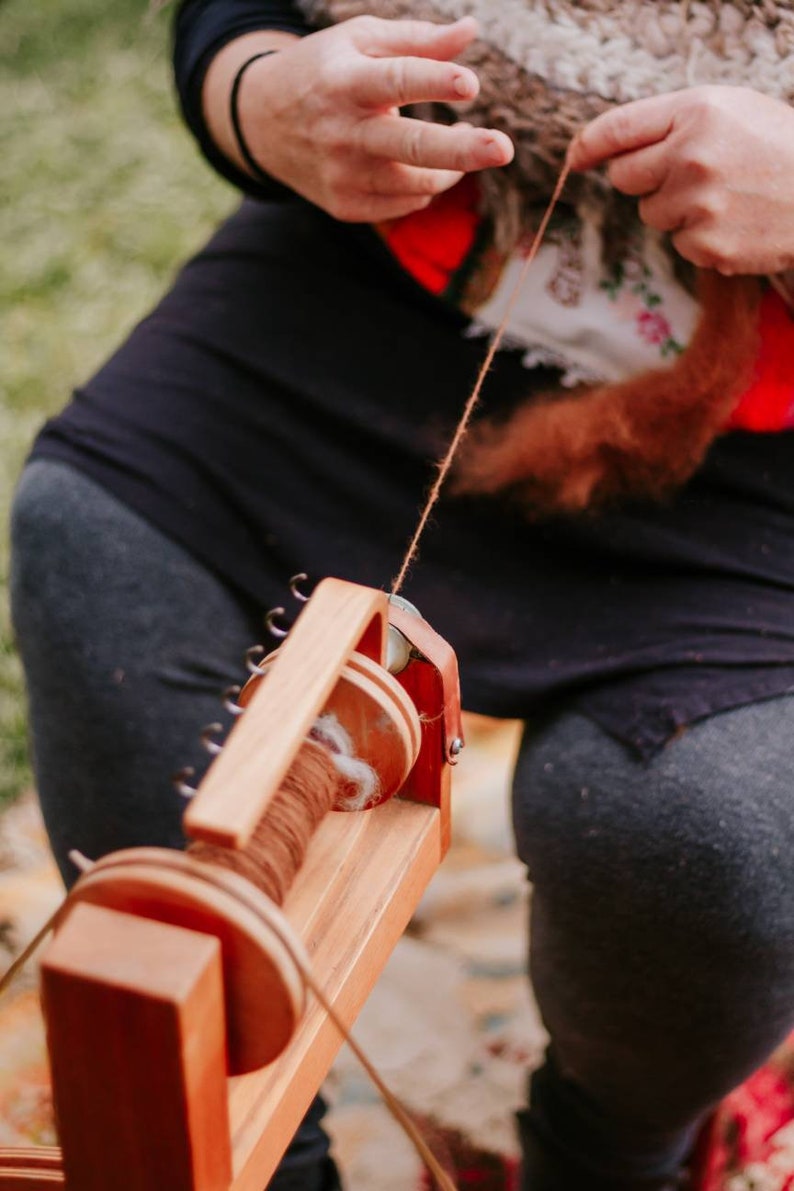 May include: A person spins brown yarn on a wooden spinning wheel. The wheel has a spool with a brown yarn and a white fluffy fiber.
