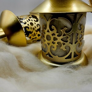 May include: Two gold metal lanterns with intricate cut-out designs. The lanterns are sitting on a white fluffy surface.