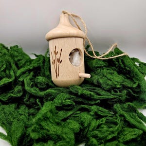 May include: A small wooden birdhouse ornament with a twig design and a hole for a bird to enter. The ornament is hanging from a twine string and is sitting on a bed of green moss.