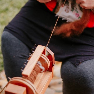 May include: A person spins brown yarn on a wooden spinning wheel. The wheel has a spool of yarn and a crank. The person is holding the yarn with their right hand and spinning the wheel with their left hand.
